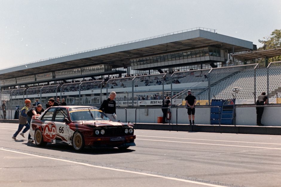 Vintage race car being pushed by crew at outdoor raceway on a sunny day.